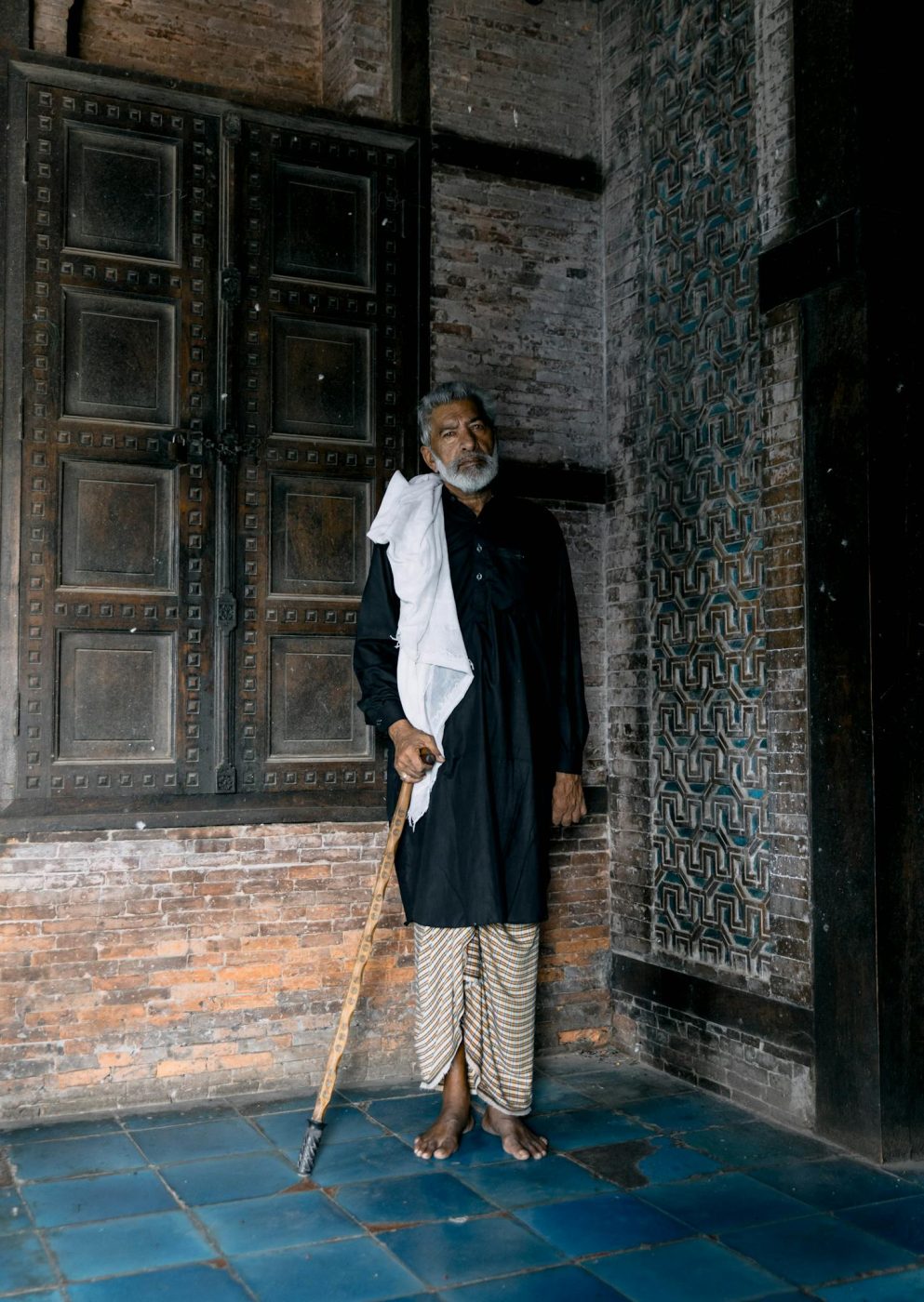 Elderly man stands against ornate wall in traditional attire, Multan, Pakistan.
