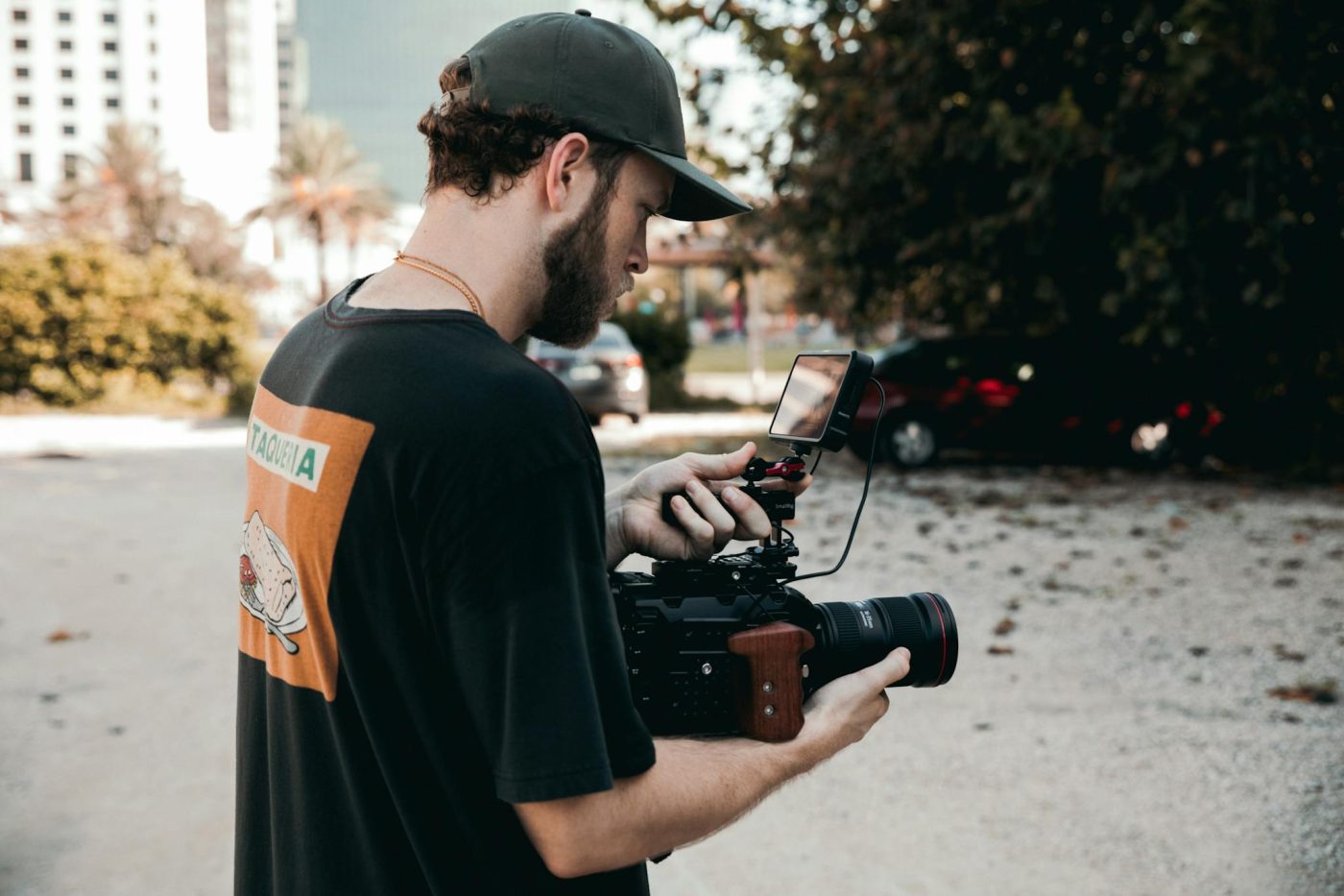 A bearded man in a cap operates a professional video camera outdoors, capturing footage.